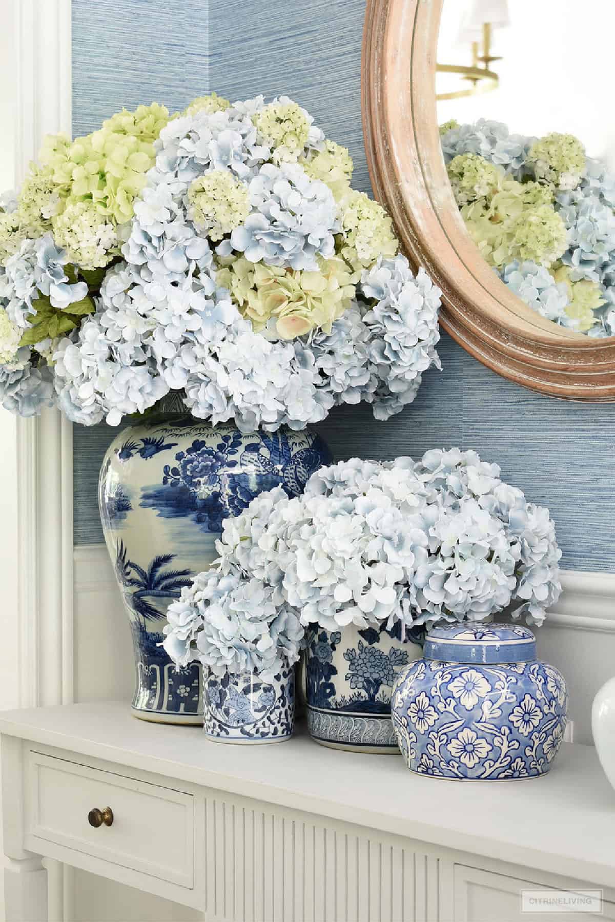 light blue and light green hydrangeas arranged in blue and white chinoiserie containers on a console table in front of a mirror