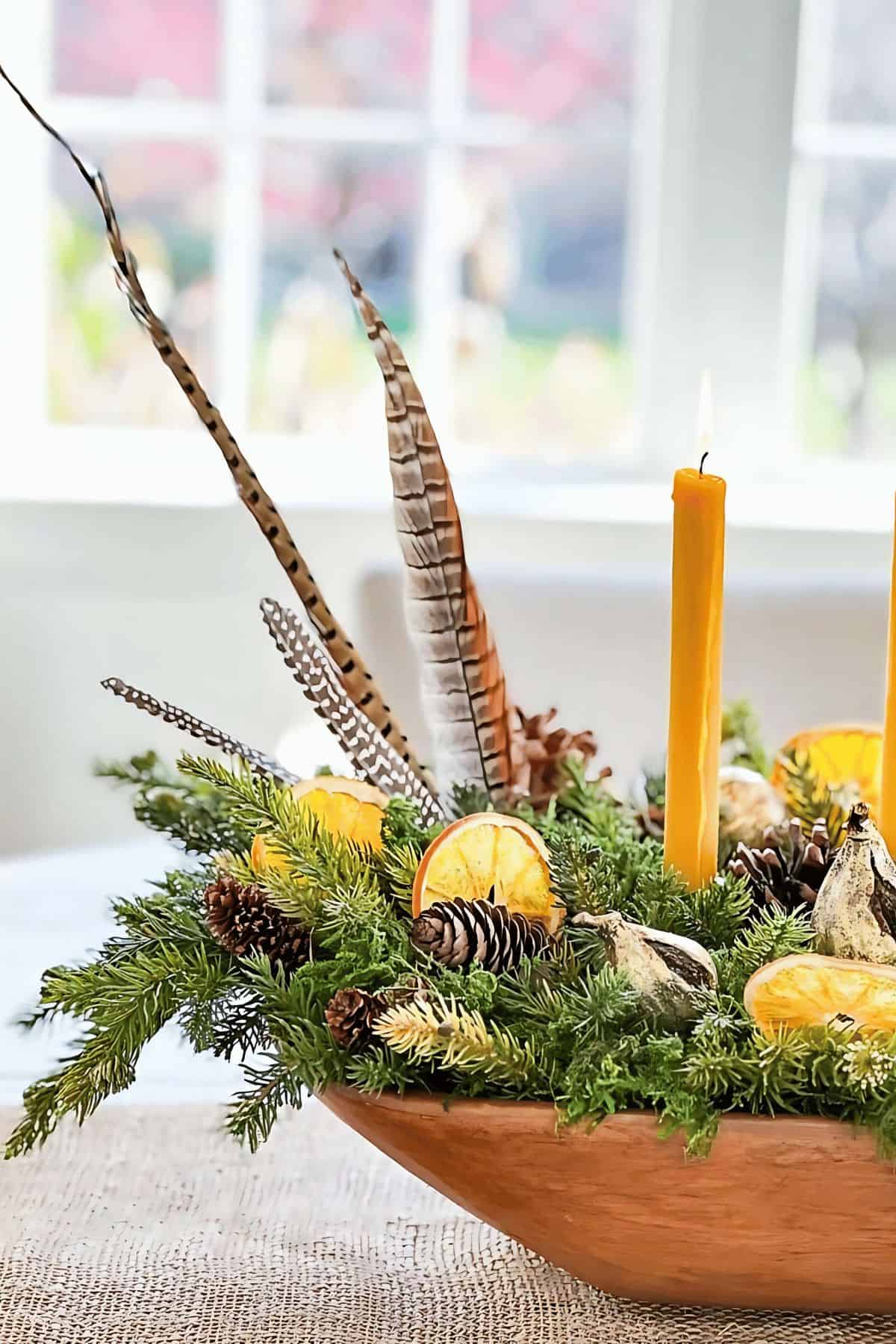 Feathered orange and pinecone centerpiece with candles and dried citrus slices for holiday decor.