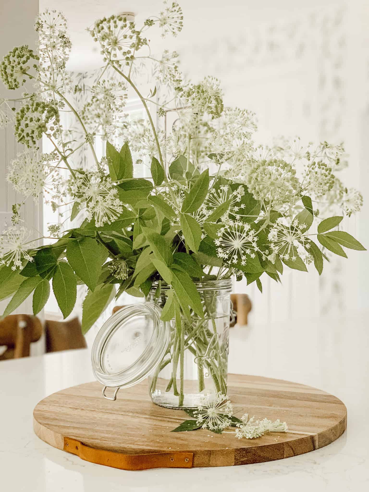glass vase on a round breadboard filled with white wild flowers and greenery