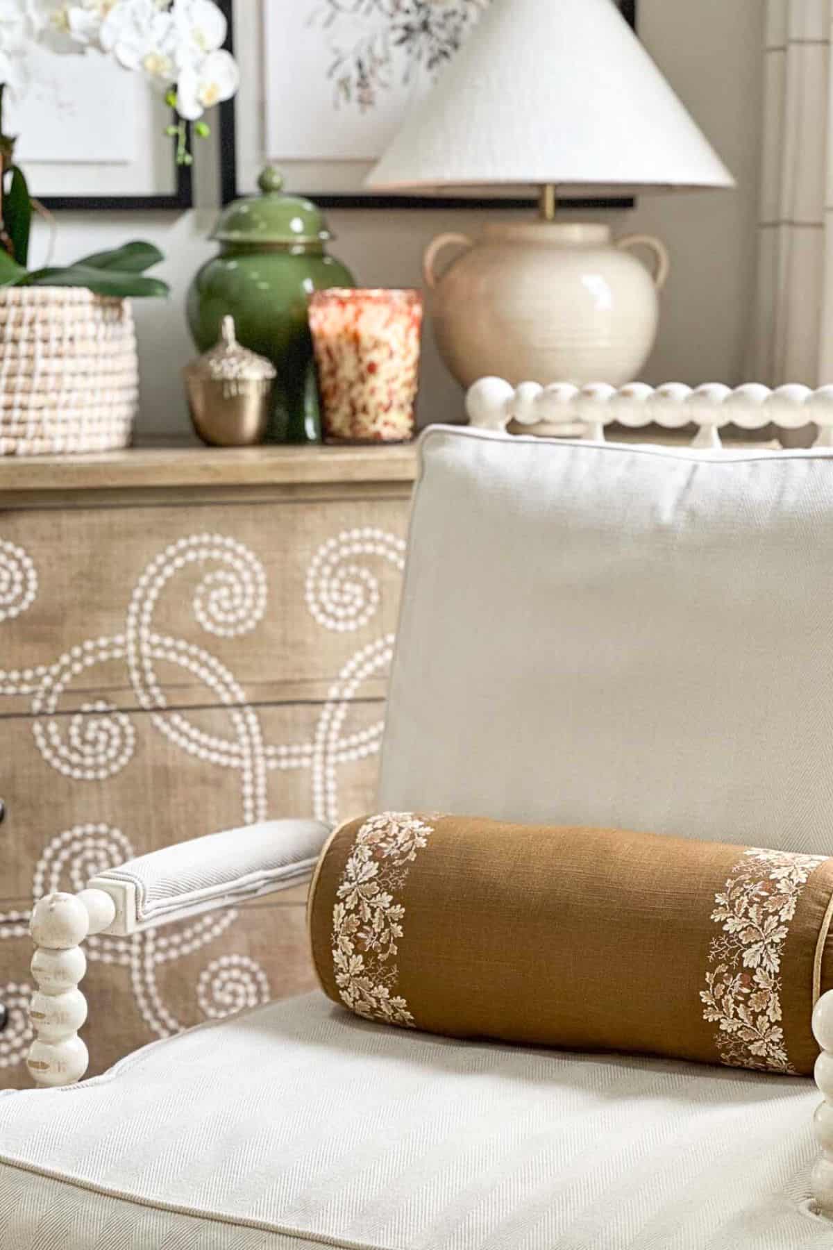 partial view of a spindle chair with cream cushions holding a brown bolster pillow with fall accessories on a chest in the background