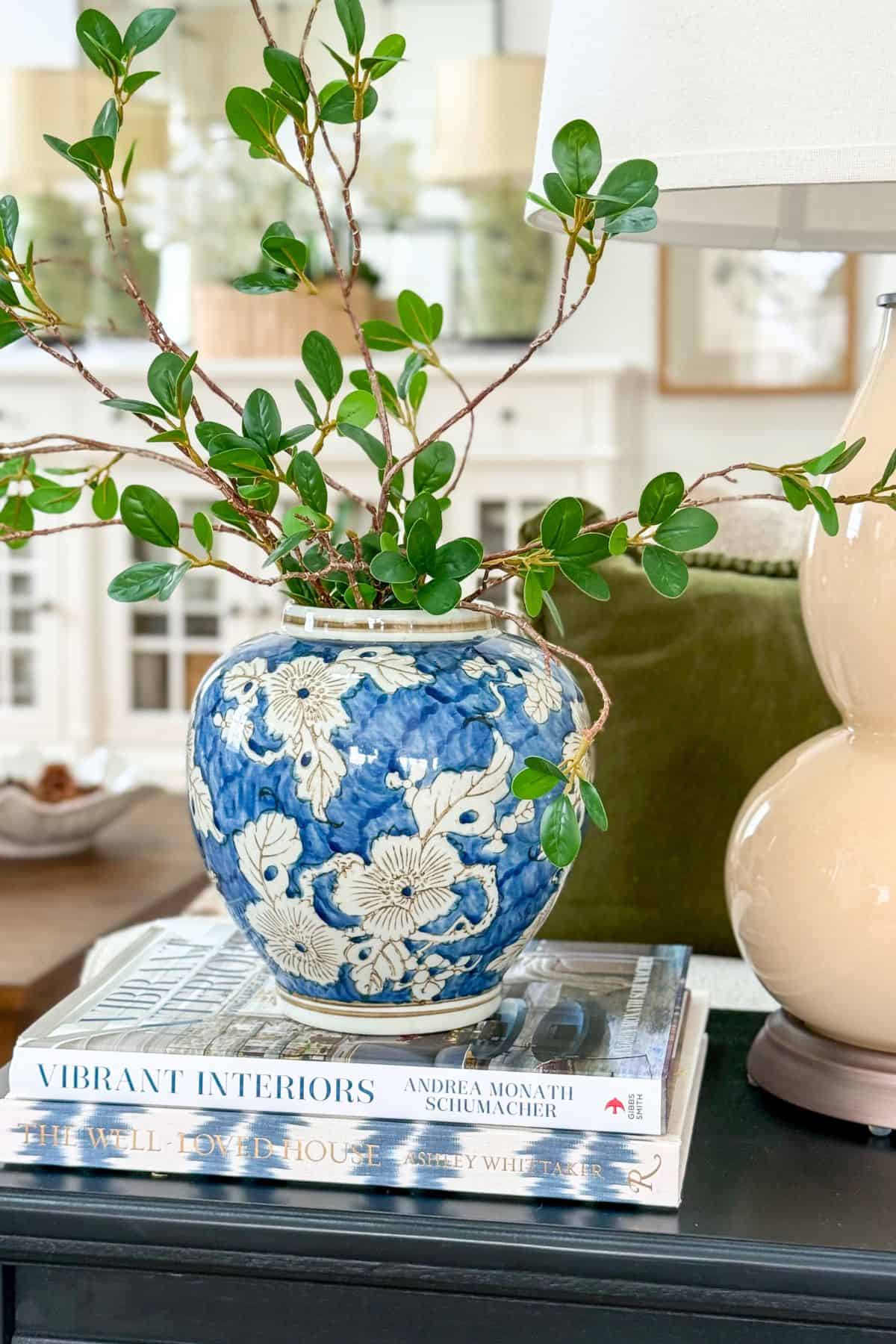 blue and white vase filled with faux greenery stems on a stack of books in a living room