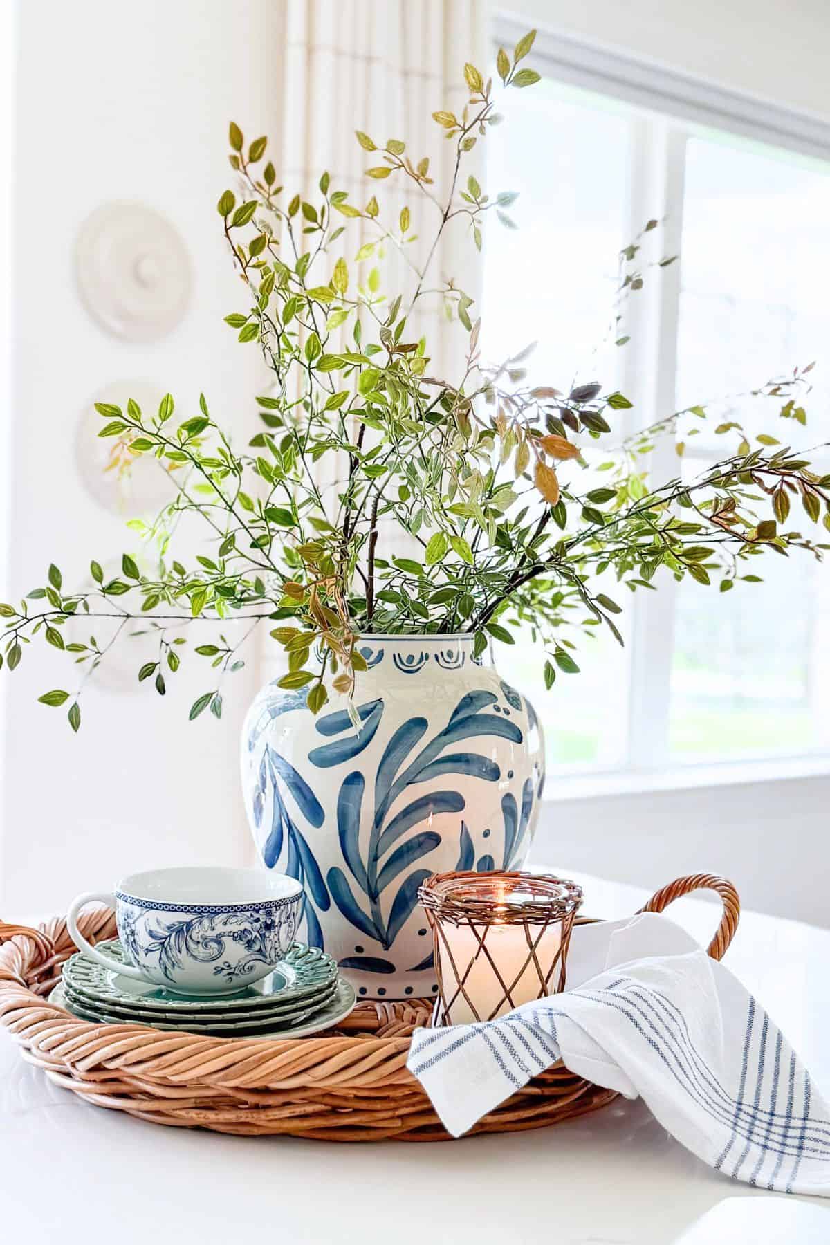 long greenery stems in a blue and white vase arranged on a round wicker tray with other accessories