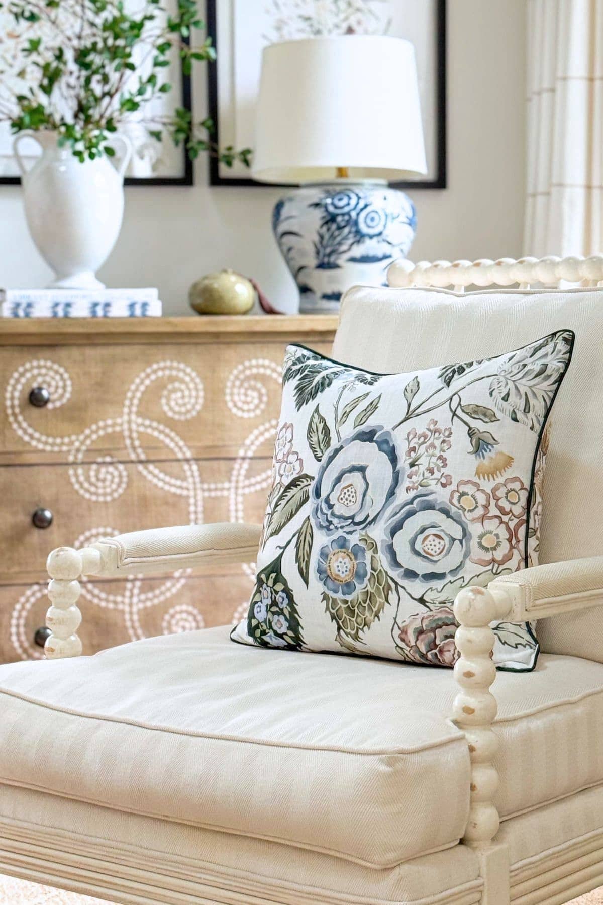 spindle chair with neutral throw pillow in front of a curlicue chest with a blue and white lamp and white vase on top