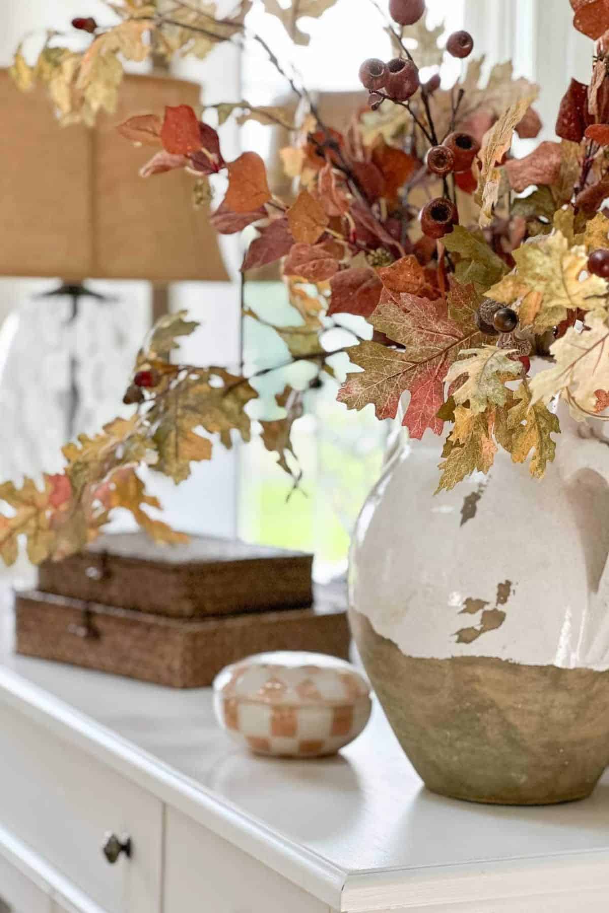 Fall arrangement with autumn leaves and berries in a rustic two-tone vase displayed on a white dresser with warm neutral accents.