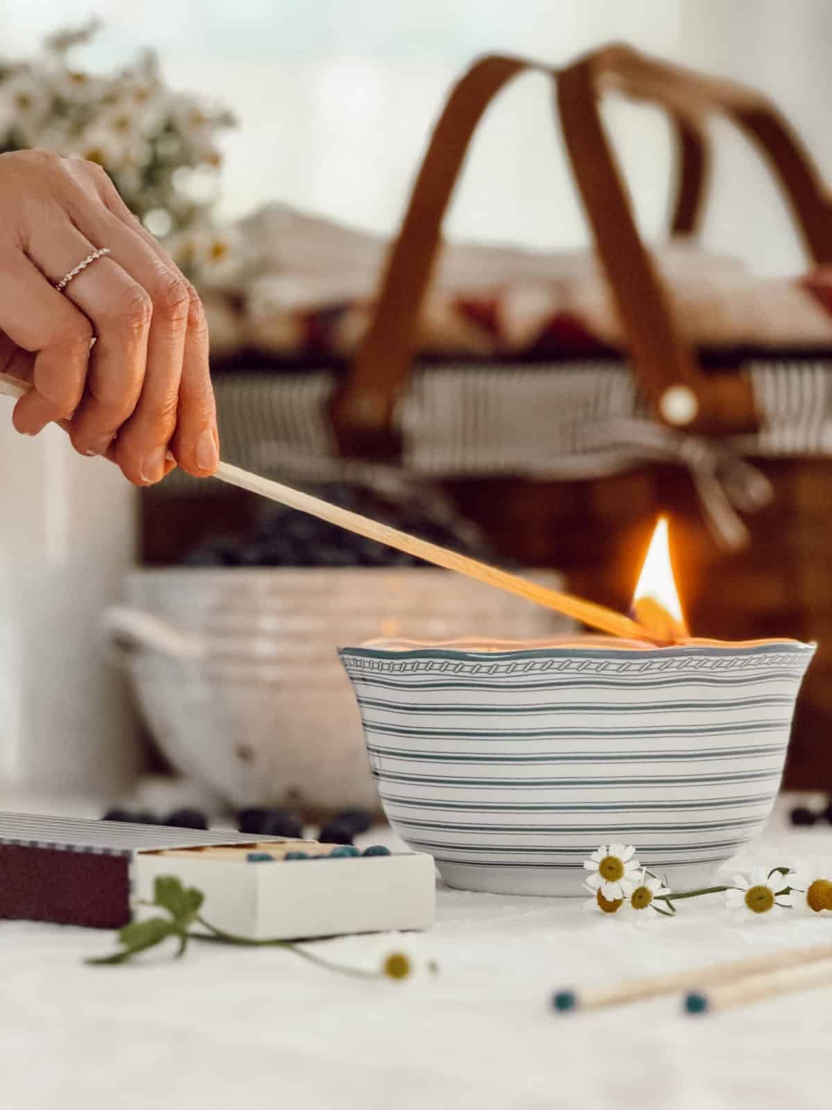 person's hand holding a long match stick to light a candle in a gray and white striped bowl