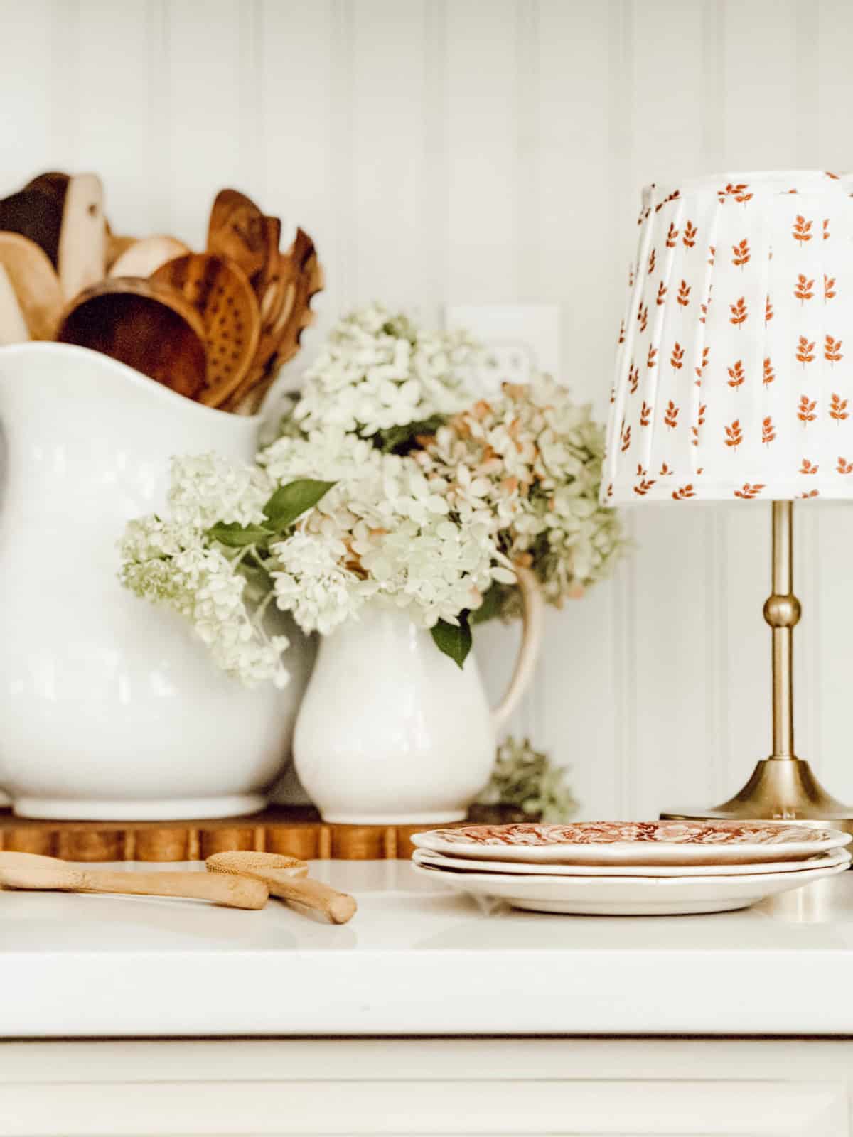 Cozy fall kitchen vignette with white hydrangeas, wooden utensils, stacked transferware plates, and a small brass lamp with a patterned shade.