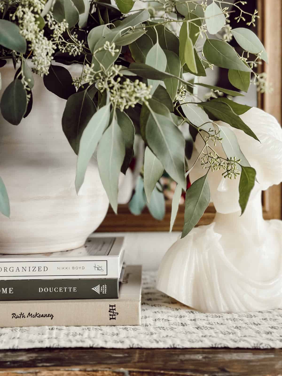 dried eucalyptus leaves in a white vase on a stack of books beside a small white bust sculpture