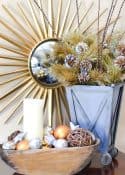 pewter container filled with gold faux pine branches and a small dough bowl filled with gold and silver ornaments in front of a starburst mirror