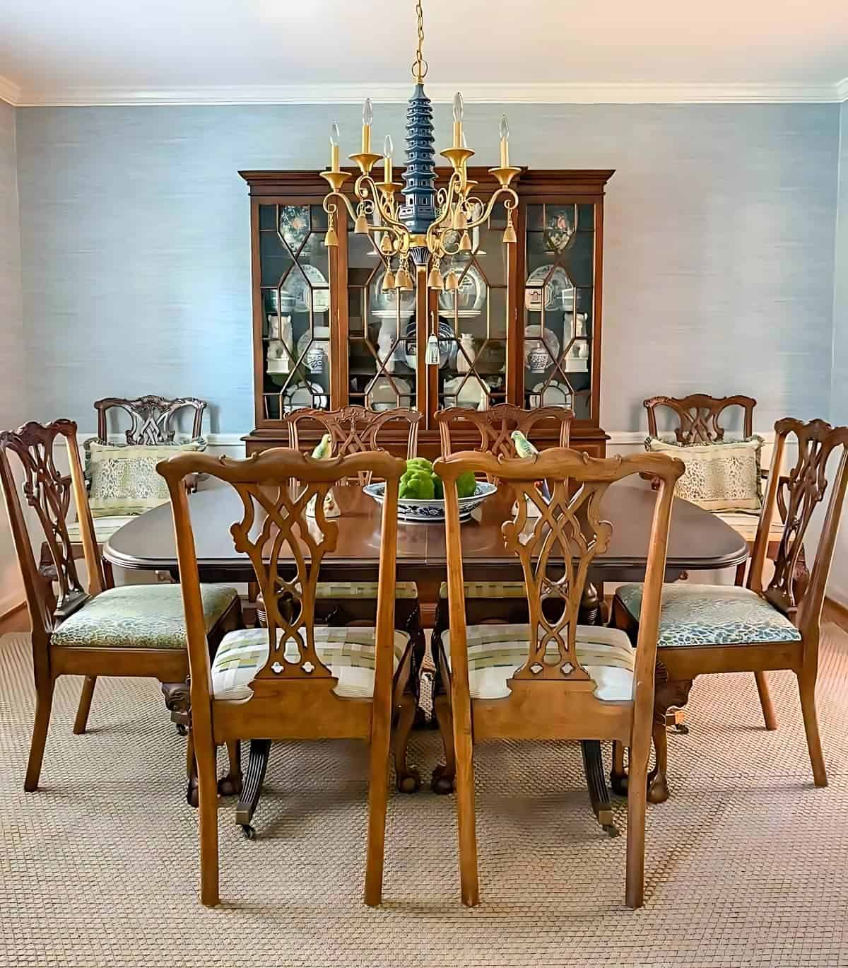 Elegant wooden dining room table with eight carved chairs and a china cabinet, illuminated by a traditional chandelier.