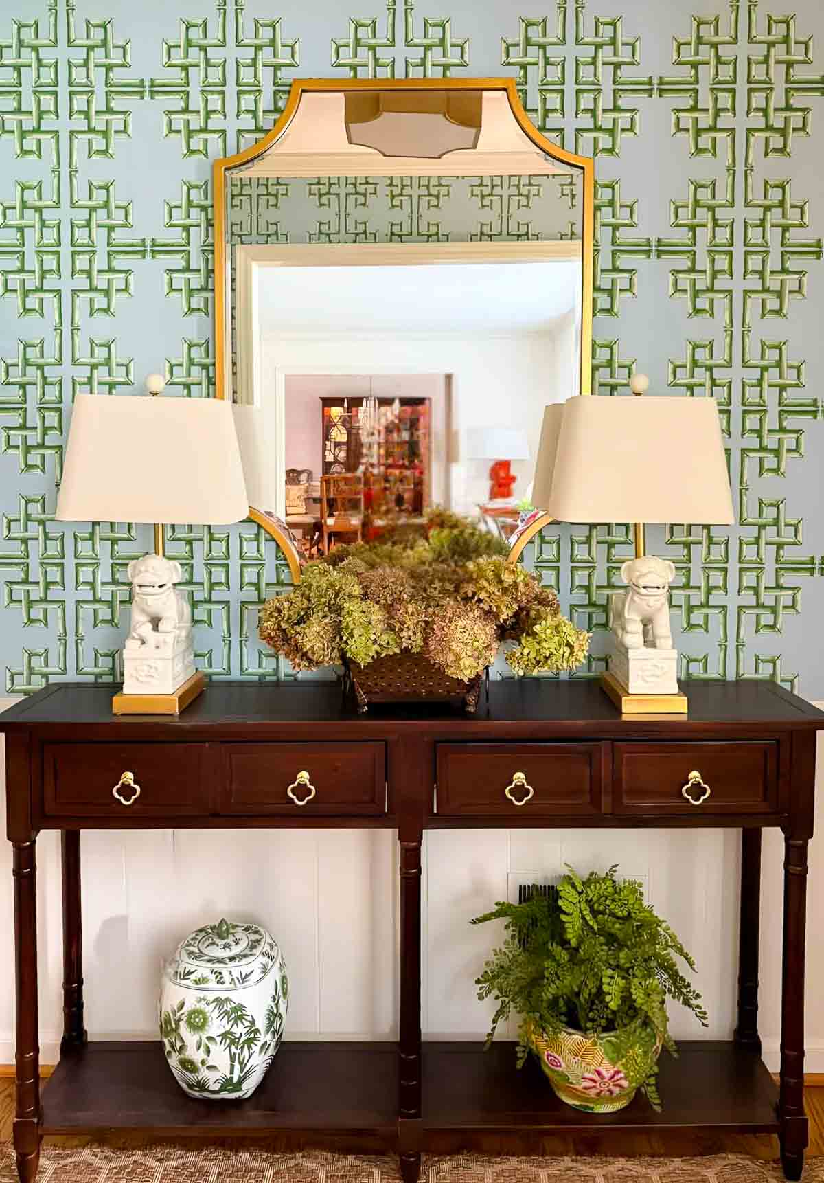 Decorative wooden console table with floral arrangements and lamps in front of intricately patterned wall.
