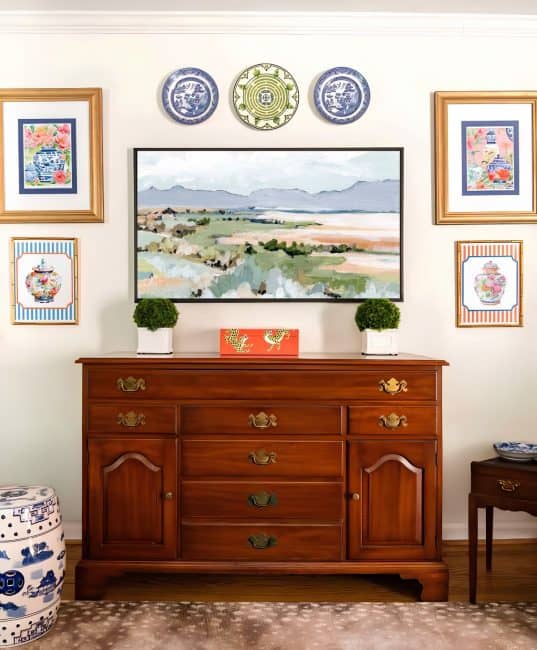 partial view of a living room decorated with colorful patterns, chinoiserie accents, and traditional style furniture. Includes a blue velvet loveseat, faux bamboo bookcase, antelope print area rug, samsung frame tv in a gallery wall with art prints, framed needlepoint and plates, with coral accents