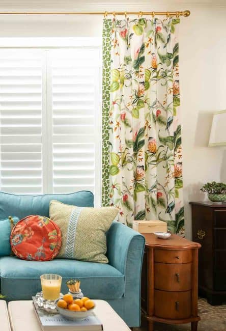 partial view of a living room decorated with colorful patterns, chinoiserie accents, and traditional style furniture. Includes a blue velvet loveseat, faux bamboo bookcase, antelope print area rug, samsung frame tv in a gallery wall with art prints, framed needlepoint and plates, with coral accents