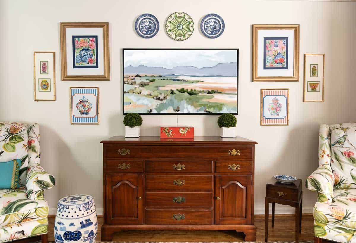 partial view of a living room decorated with colorful patterns, chinoiserie accents, and traditional style furniture. Includes a blue velvet loveseat, faux bamboo bookcase, antelope print area rug, samsung frame tv in a gallery wall with art prints, framed needlepoint and plates, with coral accents